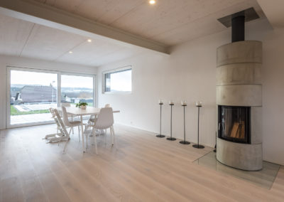 Cette image représente l'intérieur de l'espace repas d'une maison individuelle construite en ossature bois par Volery Frères SA. Les planchers sont réalisés grâce à la pose d'une dalle bois visible en plafond et blanchie. Un poêle est intégré et traverse la dalle bois.