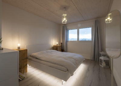 Cette image représente l'intérieur de la chambre parentale d'une maison individuelle construite en ossature bois par Volery Frères SA. Les planchers sont réalisés grâce à la pose d'une dalle bois visible en plafond et blanchie.