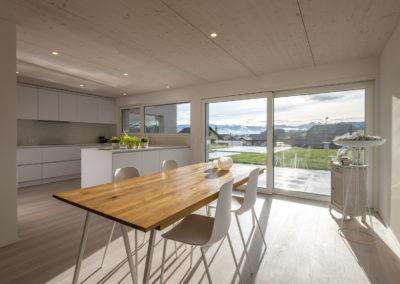 Cette image représente l'intérieur d'une maison individuelle construite en ossature bois par Volery Frères SA. Les planchers sont réalisés grâce à la pose d'une dalle bois visible en plafond et blanchie. L'espace repas/cuisine bénéficie de grande ouvertures.