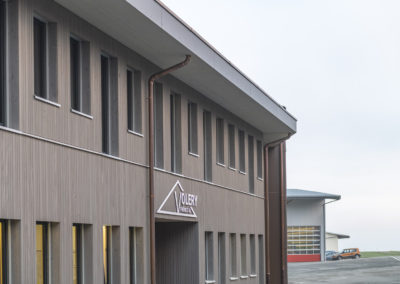 Cette image représente l'extérieur des nouveaux bureaux de l'entreprise Volery Frères SA à Aumont. Bâtiment construit en ossature bois avec revêtement en bois, il est appondu entre une halle de stockage et une halle de production de fenêtre.